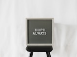 A letter board with the phrase 'Hope Always' written on it, placed on a black stool against a plain white background. The board has a light wooden frame with dark letterboard and white letters.