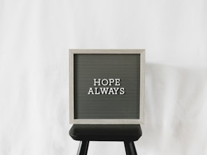 A letter board with the phrase 'Hope Always' written on it, placed on a black stool against a plain white background. The board has a light wooden frame with dark letterboard and white letters.