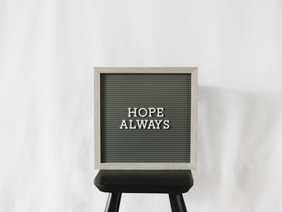 A letter board with the phrase 'Hope Always' written on it, placed on a black stool against a plain white background. The board has a light wooden frame with dark letterboard and white letters.