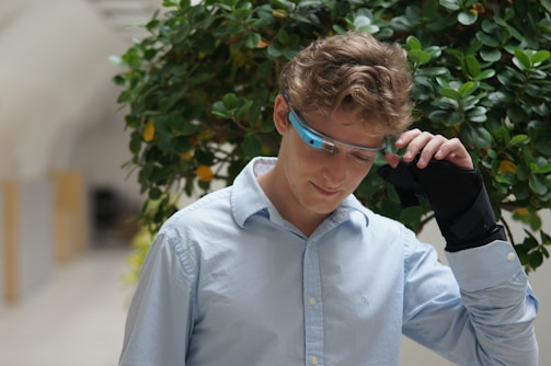 A person wearing a light blue shirt is adjusting a pair of wearable smart glasses. The background features lush green foliage with some yellow accents. The person appears to be thoughtful, with eyes looking downward and a slight smile.