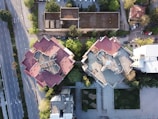 Aerial view of two large, adjacent buildings with red roofs, situated in an urban environment. The buildings are surrounded by trees and a network of roads, with several cars parked nearby. The image captures a blend of architecture and greenery, highlighting the contrast between human-made structures and natural elements.