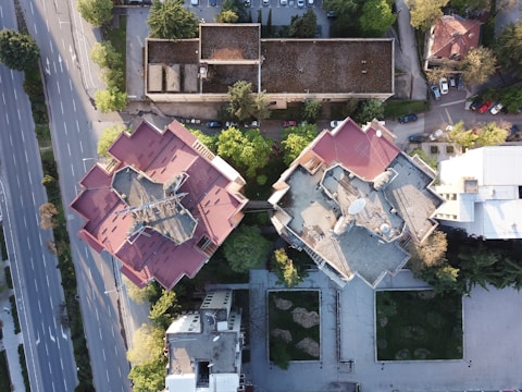 Aerial view of two large, adjacent buildings with red roofs, situated in an urban environment. The buildings are surrounded by trees and a network of roads, with several cars parked nearby. The image captures a blend of architecture and greenery, highlighting the contrast between human-made structures and natural elements.