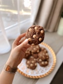 A hand wearing a bracelet is holding a chocolate cookie with white chocolate chips, set against a blurred backdrop of a stack of similar cookies on a decorative plate. Soft light streams through sheer curtains in the background.