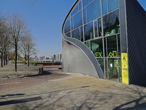 A modern building with large glass windows and an architectural design characterized by curved lines. The exterior features a combination of metal and glass materials, and there are yellow accents around the entrance. Leafless trees and a paved walkway are in the vicinity, and the background reveals a cityscape under a clear blue sky.