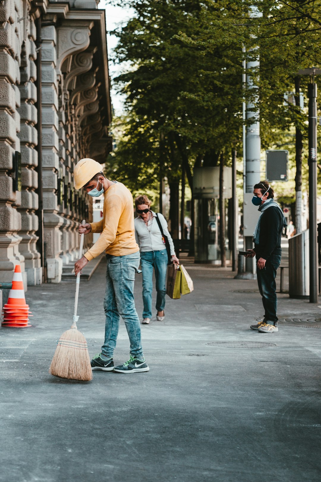 man in blue dress shirt and yellow pants holding broom while walking on sidewalk during daytime