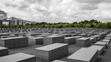 An array of large, rectangular concrete blocks is arranged in a grid pattern, creating a geometric landscape. These blocks are situated on a paved ground, with a backdrop of green trees and a cloudy sky overhead. Buildings are visible in the distance, contrasting with the solemn atmosphere of the rows of blocks.