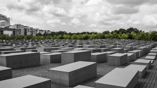 An array of large, rectangular concrete blocks is arranged in a grid pattern, creating a geometric landscape. These blocks are situated on a paved ground, with a backdrop of green trees and a cloudy sky overhead. Buildings are visible in the distance, contrasting with the solemn atmosphere of the rows of blocks.