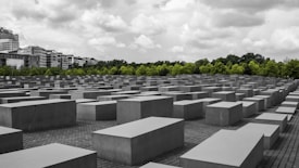 An array of large, rectangular concrete blocks is arranged in a grid pattern, creating a geometric landscape. These blocks are situated on a paved ground, with a backdrop of green trees and a cloudy sky overhead. Buildings are visible in the distance, contrasting with the solemn atmosphere of the rows of blocks.