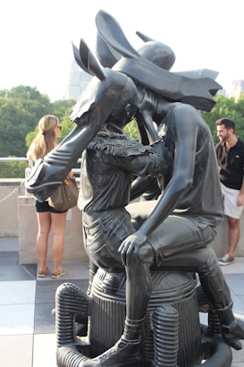 A black sculpture of two figures sitting closely together, with one person wearing a large hat and the other having a donkey head. The sculpture is placed on a rooftop terrace with trees and city buildings visible in the background. There are people casually standing nearby observing the artwork.