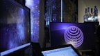 Several computer screens are displaying digital art and animations, including swirling galactic visuals and geometric designs. The atmosphere is futuristic and artistic, enhanced by ambient lighting. Various objects such as bottles and figurines appear in the background, creating an eclectic and creative environment.