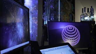 Several computer screens are displaying digital art and animations, including swirling galactic visuals and geometric designs. The atmosphere is futuristic and artistic, enhanced by ambient lighting. Various objects such as bottles and figurines appear in the background, creating an eclectic and creative environment.