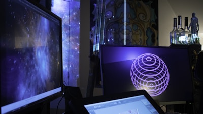 Several computer screens are displaying digital art and animations, including swirling galactic visuals and geometric designs. The atmosphere is futuristic and artistic, enhanced by ambient lighting. Various objects such as bottles and figurines appear in the background, creating an eclectic and creative environment.