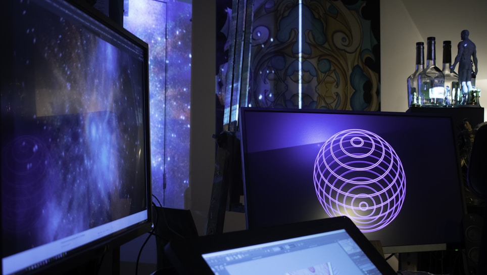 Several computer screens are displaying digital art and animations, including swirling galactic visuals and geometric designs. The atmosphere is futuristic and artistic, enhanced by ambient lighting. Various objects such as bottles and figurines appear in the background, creating an eclectic and creative environment.