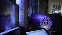Several computer screens are displaying digital art and animations, including swirling galactic visuals and geometric designs. The atmosphere is futuristic and artistic, enhanced by ambient lighting. Various objects such as bottles and figurines appear in the background, creating an eclectic and creative environment.
