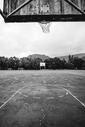 An abandoned basketball court with a weathered backboard and hoop. The cracked cement surface stretches out with faint court lines barely visible. In the background, there are two empty soccer goals surrounded by dense trees and mountains under a cloudy sky.