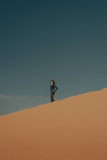 A person stands confidently on a sand dune under a clear blue sky, with the wind slightly blowing, creating a serene and solitary atmosphere.