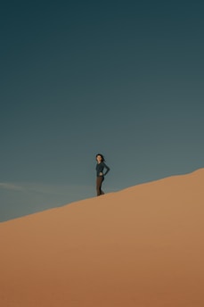 A person stands confidently on a sand dune under a clear blue sky, with the wind slightly blowing, creating a serene and solitary atmosphere.
