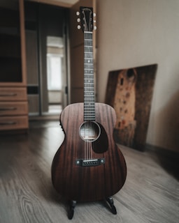 An acoustic guitar with a rich, dark wood finish is placed on a stand in a room with wooden flooring. In the background, there is a blurred image of a partially visible painting and furniture, suggesting a cozy indoor setting.