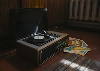 A vintage record player with a vinyl disc spinning on it, surrounded by a stack of vinyl record covers on a wooden floor. The setting appears to be indoors, with dim lighting coming from a nearby window. A radiator is visible in the background, adding to the nostalgic and retro ambiance.