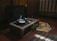 A vintage record player with a vinyl disc spinning on it, surrounded by a stack of vinyl record covers on a wooden floor. The setting appears to be indoors, with dim lighting coming from a nearby window. A radiator is visible in the background, adding to the nostalgic and retro ambiance.