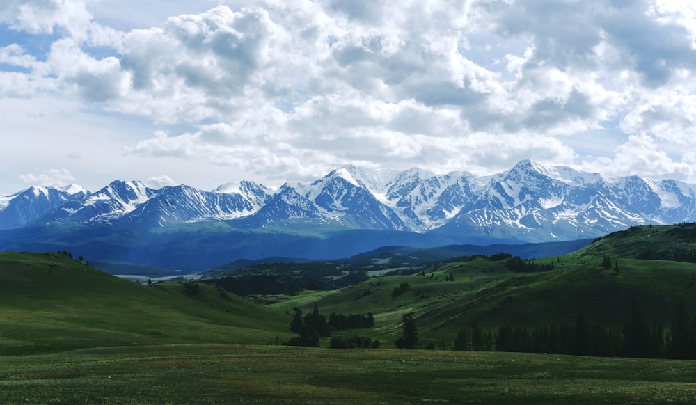 green grass field near snow covered mountains under white clouds and blue sky during daytime