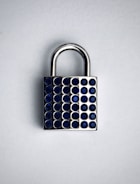 A silver padlock with a rectangular body and a curved shackle sits against a light background. The front of the padlock is adorned with rows of small, circular blue gems, giving it a decorative appearance.