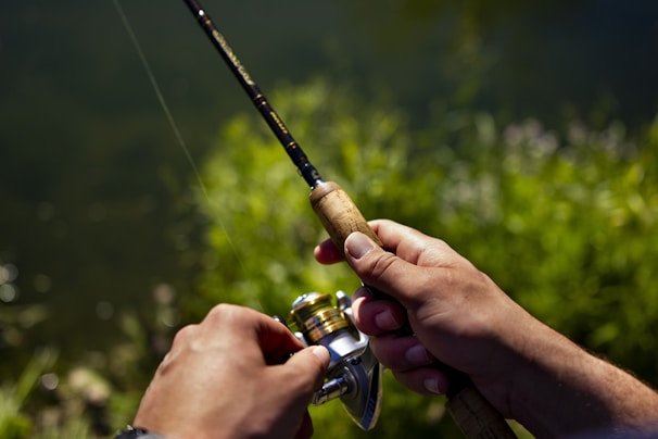 Hands holding a fishing rod with a wooden grip, situated near a body of water and surrounded by greenery, indicating an outdoor fishing scene.