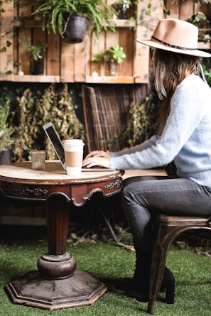 A person wearing a hat and a light blue sweater is sitting at a round wooden table with a laptop. A coffee cup and a candle are also on the table. The setting includes indoor plants hanging on the wall and a floor covered with artificial grass, creating a cozy and nature-inspired atmosphere.
