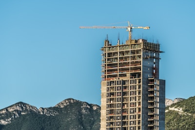 A tall building under construction with a crane on top, set against a clear blue sky. In the background, there are green, rugged mountains providing a dramatic backdrop to the urban development.