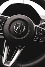 A close-up view of a car steering wheel with a logo prominently displayed in the center. The steering wheel is black with metallic accents, and various control buttons are visible on the sides.