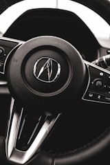 A close-up view of a car steering wheel with a logo prominently displayed in the center. The steering wheel is black with metallic accents, and various control buttons are visible on the sides.