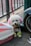 A small poodle with distinct fluffy white fur is sitting on a pavement, adorned with pink ears and green paws. The dog is leashed, next to a pink and white plastic container, by a railing and a scooter tire. A silver bell hangs from its red collar.