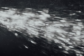 A dynamic burst of light particles streaks across a dark background, creating an abstract and energetic visual effect. The particles appear to be moving at high speed, with varying intensities and sizes, giving the image a sense of motion and depth.