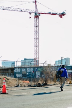 A security guard wearing a blue jacket walks along a fenced construction site with graffiti on the walls. A large red and white construction crane towers in the background, with several high-rise buildings visible in the distance. The area has some traffic cones placed sporadically and appears deserted aside from the single person.
