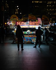 A street food cart illuminated at night with a bright sign advertising drinks and snacks. Four people are gathered around the cart, which is covered with branded umbrellas. The scene is set in an urban environment, with the glow of city lights and the silhouettes of tall buildings in the background.