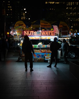 A street food cart illuminated at night with a bright sign advertising drinks and snacks. Four people are gathered around the cart, which is covered with branded umbrellas. The scene is set in an urban environment, with the glow of city lights and the silhouettes of tall buildings in the background.