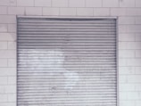 A metal roll-up shutter door is closed against a tiled wall. The door features horizontal slats and shows signs of wear and weathering. The wall consists of rectangular white tiles with a slightly worn appearance.