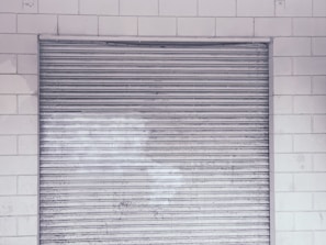 A metal roll-up shutter door is closed against a tiled wall. The door features horizontal slats and shows signs of wear and weathering. The wall consists of rectangular white tiles with a slightly worn appearance.