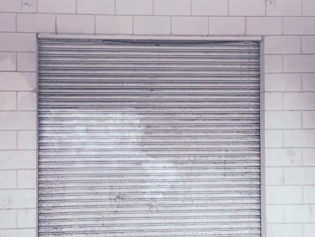 Technician repairing a metal shutter on a storefront with tools in hand.