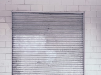 A metal roll-up shutter door is closed against a tiled wall. The door features horizontal slats and shows signs of wear and weathering. The wall consists of rectangular white tiles with a slightly worn appearance.