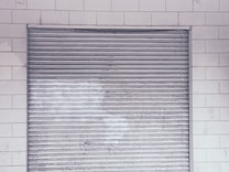 A metal roll-up shutter door is closed against a tiled wall. The door features horizontal slats and shows signs of wear and weathering. The wall consists of rectangular white tiles with a slightly worn appearance.