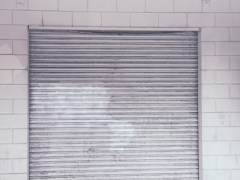 A metal roll-up shutter door is closed against a tiled wall. The door features horizontal slats and shows signs of wear and weathering. The wall consists of rectangular white tiles with a slightly worn appearance.
