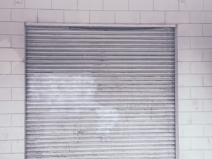 A metal roll-up shutter door is closed against a tiled wall. The door features horizontal slats and shows signs of wear and weathering. The wall consists of rectangular white tiles with a slightly worn appearance.