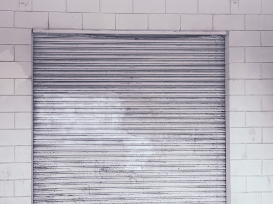 A metal roll-up shutter door is closed against a tiled wall. The door features horizontal slats and shows signs of wear and weathering. The wall consists of rectangular white tiles with a slightly worn appearance.