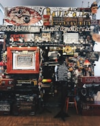 A dense collage of text, objects, and various art pieces displayed on a wall. The composition involves handwritten and printed text in different languages with a variety of fonts and sizes. There are numerous items like a red circular sign, painted boards, various images, a small lamp, and a wooden chair. The art appears eclectic and chaotic, with elements overlapping and layered.
