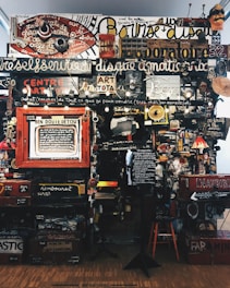 A dense collage of text, objects, and various art pieces displayed on a wall. The composition involves handwritten and printed text in different languages with a variety of fonts and sizes. There are numerous items like a red circular sign, painted boards, various images, a small lamp, and a wooden chair. The art appears eclectic and chaotic, with elements overlapping and layered.