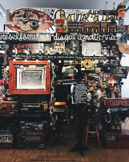 A dense collage of text, objects, and various art pieces displayed on a wall. The composition involves handwritten and printed text in different languages with a variety of fonts and sizes. There are numerous items like a red circular sign, painted boards, various images, a small lamp, and a wooden chair. The art appears eclectic and chaotic, with elements overlapping and layered.