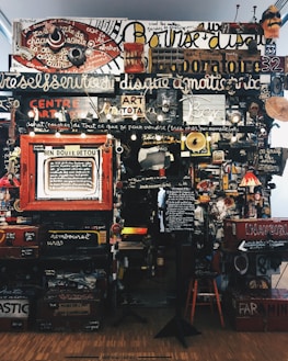 A dense collage of text, objects, and various art pieces displayed on a wall. The composition involves handwritten and printed text in different languages with a variety of fonts and sizes. There are numerous items like a red circular sign, painted boards, various images, a small lamp, and a wooden chair. The art appears eclectic and chaotic, with elements overlapping and layered.