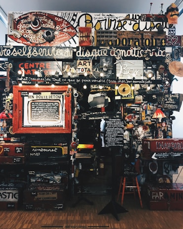 A dense collage of text, objects, and various art pieces displayed on a wall. The composition involves handwritten and printed text in different languages with a variety of fonts and sizes. There are numerous items like a red circular sign, painted boards, various images, a small lamp, and a wooden chair. The art appears eclectic and chaotic, with elements overlapping and layered.