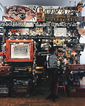 A dense collage of text, objects, and various art pieces displayed on a wall. The composition involves handwritten and printed text in different languages with a variety of fonts and sizes. There are numerous items like a red circular sign, painted boards, various images, a small lamp, and a wooden chair. The art appears eclectic and chaotic, with elements overlapping and layered.